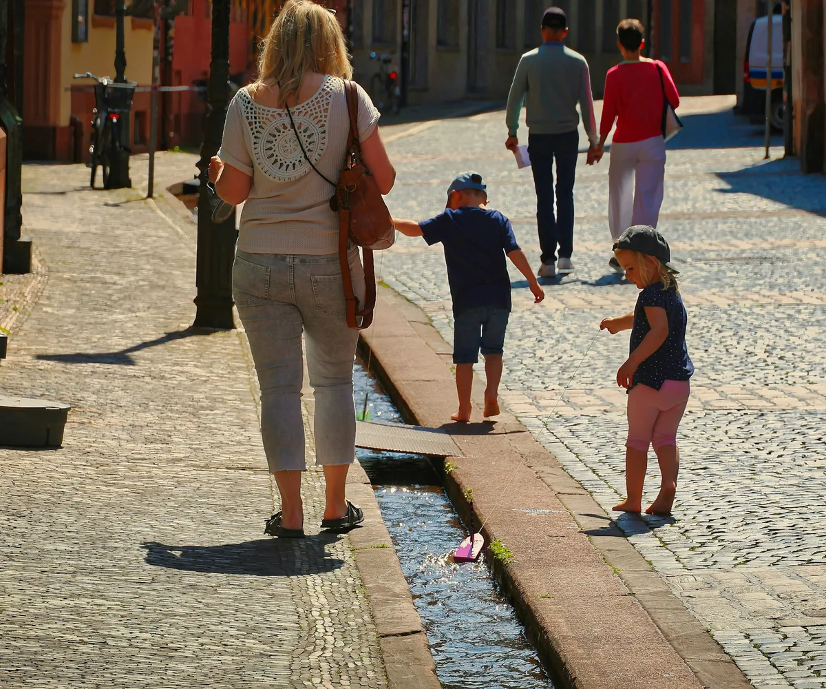 Parents swing their child between them as they walk a sunlit cobblestone street in a European old town